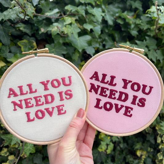 Hand holding two hand embroidery hoops with pink and white fabric and pink typography stitching saying All you need is love