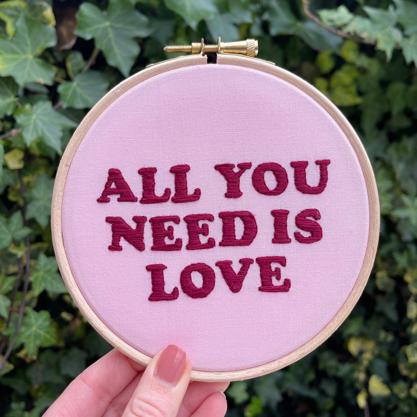 Embroidery hoop with pink fabric and red hand embroidered typography lettering quote saying 'all you need is love'